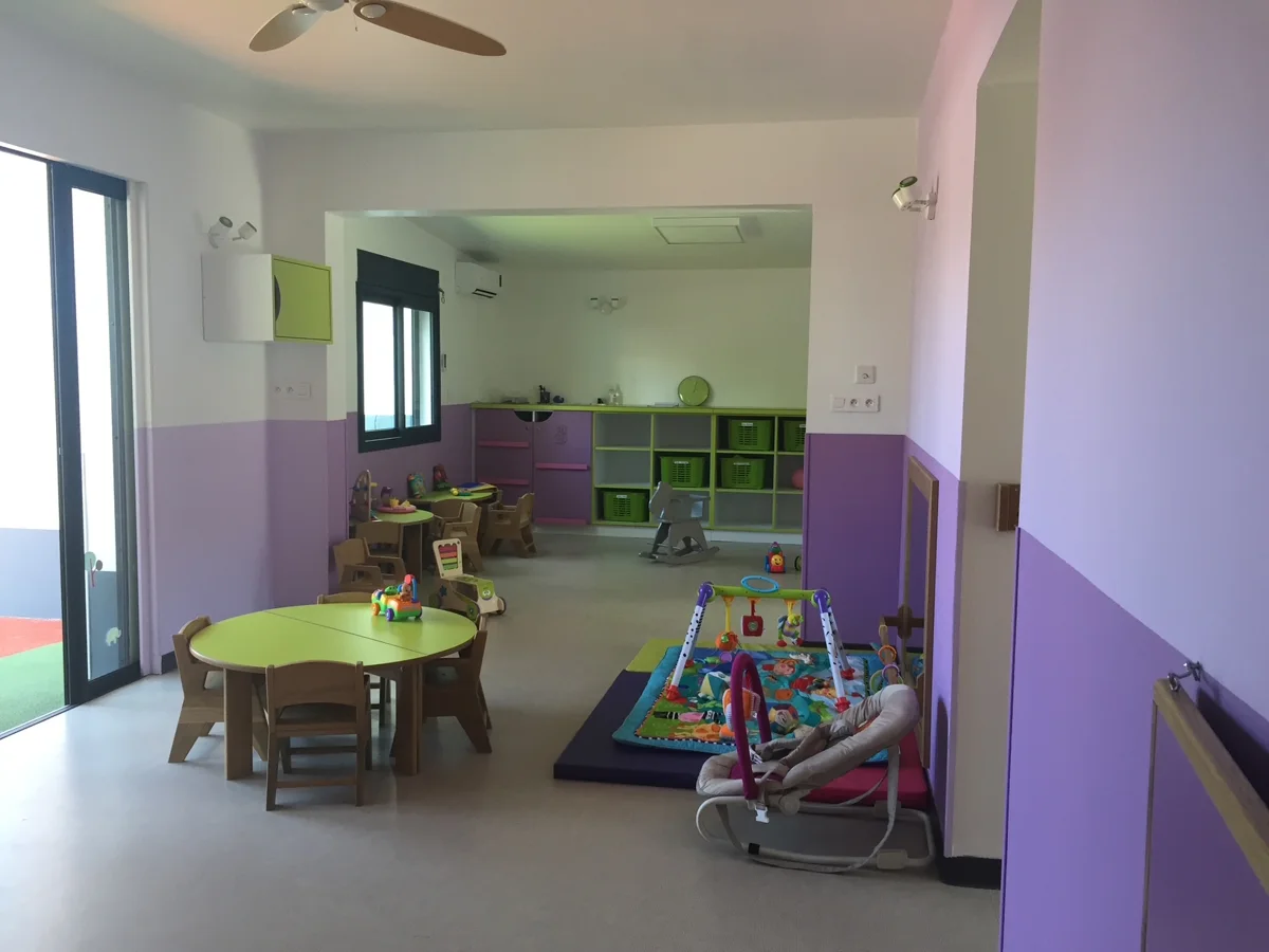 Salle de vie lumineuse d'une crèche aux murs violets et blancs, avec table ronde verte, petites chaises en bois, rangements et tapis d'éveil ; idéale pour présenter les espaces d'accueil.
